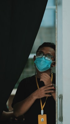 A person wearing glasses and a blue surgical mask stands near an open door with a dark curtain partially hiding them. They are looking upwards with their hands raised near their chest. A yellow lanyard with an ID card hangs around their neck.