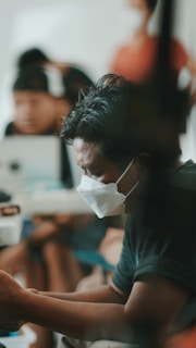 A person is wearing a face mask, appearing to be engrossed in their activity. The background is softly blurred, with other individuals present, possibly in a waiting room or similar setting. The image conveys a sense of focus and attentiveness.