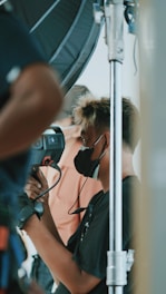 A person wearing a black face mask is operating a professional video camera on a set. The scene involves technical equipment and another person is partially visible in the background, likely involved in the filming process.