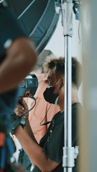 A person wearing a black face mask is operating a professional video camera on a set. The scene involves technical equipment and another person is partially visible in the background, likely involved in the filming process.