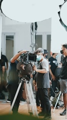 A group of people, some wearing masks, are gathered around a professional camera setup on a tripod. The setting appears to be a film or video shoot, with bright studio lights and equipment visible in the background.