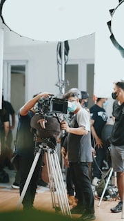 A group of people, some wearing masks, are gathered around a professional camera setup on a tripod. The setting appears to be a film or video shoot, with bright studio lights and equipment visible in the background.
