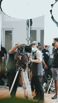A group of people, some wearing masks, are gathered around a professional camera setup on a tripod. The setting appears to be a film or video shoot, with bright studio lights and equipment visible in the background.