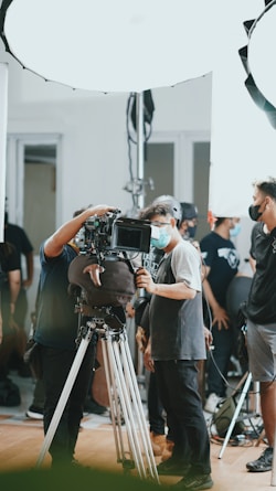 A group of people, some wearing masks, are gathered around a professional camera setup on a tripod. The setting appears to be a film or video shoot, with bright studio lights and equipment visible in the background.