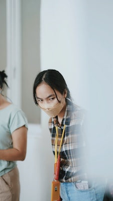 A person wearing a plaid shirt and a face mask looks intently while holding an ID badge and lanyard. The background appears blurred, and another person partly visible on the left is wearing a light-colored shirt.