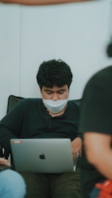 A person with dark hair and a facial mask is seated, focused on a laptop. The setting appears informal, as the individual is leaning back in a black shirt. There are blurred figures in the foreground and background, suggesting a casual or relaxed environment.