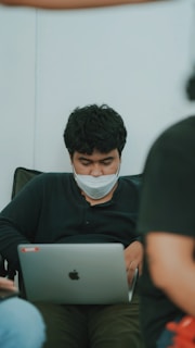 A person with dark hair and a facial mask is seated, focused on a laptop. The setting appears informal, as the individual is leaning back in a black shirt. There are blurred figures in the foreground and background, suggesting a casual or relaxed environment.