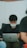 A person with dark hair and a facial mask is seated, focused on a laptop. The setting appears informal, as the individual is leaning back in a black shirt. There are blurred figures in the foreground and background, suggesting a casual or relaxed environment.