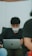A person with dark hair and a facial mask is seated, focused on a laptop. The setting appears informal, as the individual is leaning back in a black shirt. There are blurred figures in the foreground and background, suggesting a casual or relaxed environment.