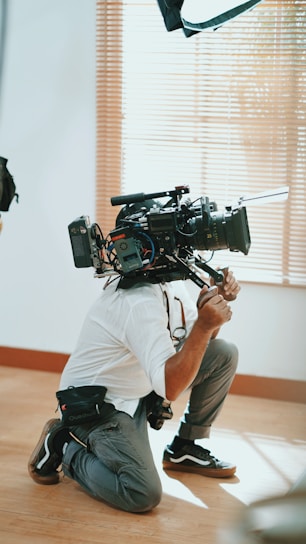 A person kneels on the floor holding a large professional video camera. The setting appears to be indoors with light wooden flooring and vertical blinds partially open in the background. The camera is equipped with various attachments and cables.