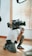 A person kneels on the floor holding a large professional video camera. The setting appears to be indoors with light wooden flooring and vertical blinds partially open in the background. The camera is equipped with various attachments and cables.
