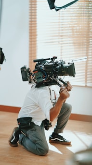 A person kneels on the floor holding a large professional video camera. The setting appears to be indoors with light wooden flooring and vertical blinds partially open in the background. The camera is equipped with various attachments and cables.