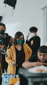 A group of people in a room, some wearing face masks. One person in a yellow jacket holds a phone, while another person interacts with equipment. There are bottles of water on a table.