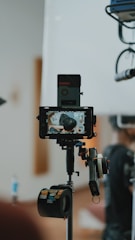 A professional camera setup is in the foreground, capturing a scene. The camera is mounted on a tripod with various accessories attached, including a monitor displaying the live capture of a person from behind. The background is blurred, focusing on the equipment.