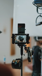 A professional camera setup is in the foreground, capturing a scene. The camera is mounted on a tripod with various accessories attached, including a monitor displaying the live capture of a person from behind. The background is blurred, focusing on the equipment.