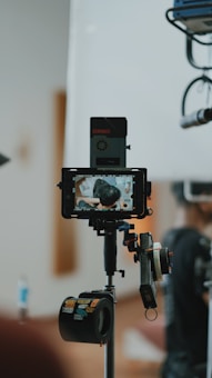 A professional camera setup is in the foreground, capturing a scene. The camera is mounted on a tripod with various accessories attached, including a monitor displaying the live capture of a person from behind. The background is blurred, focusing on the equipment.