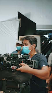 A person wearing a blue surgical mask is handling a large, professional video camera. There are other individuals in the background, also wearing masks, suggesting a film set. The setting includes equipment and backdrops commonly found in a studio environment.