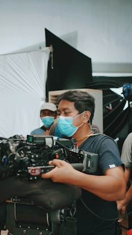 A person wearing a blue surgical mask is handling a large, professional video camera. There are other individuals in the background, also wearing masks, suggesting a film set. The setting includes equipment and backdrops commonly found in a studio environment.
