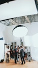 A group of people are gathered in a room, surrounded by photography or film equipment such as lights, tripods, and softboxes. Some individuals appear to be adjusting a camera on a tripod while others are engaged in conversation or checking equipment. The setting appears to be a professional studio environment with a high ceiling and white walls.