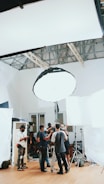 A group of people are gathered in a room, surrounded by photography or film equipment such as lights, tripods, and softboxes. Some individuals appear to be adjusting a camera on a tripod while others are engaged in conversation or checking equipment. The setting appears to be a professional studio environment with a high ceiling and white walls.