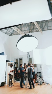 A group of people are gathered in a room, surrounded by photography or film equipment such as lights, tripods, and softboxes. Some individuals appear to be adjusting a camera on a tripod while others are engaged in conversation or checking equipment. The setting appears to be a professional studio environment with a high ceiling and white walls.