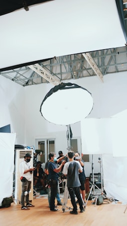 A group of people are gathered in a room, surrounded by photography or film equipment such as lights, tripods, and softboxes. Some individuals appear to be adjusting a camera on a tripod while others are engaged in conversation or checking equipment. The setting appears to be a professional studio environment with a high ceiling and white walls.