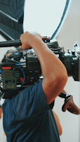 A person operates a large professional camera, holding it on their shoulder. The camera is equipped with various controls, cables, and a matte box. Strong lighting is visible in the background, indicating a studio or professional filming environment.