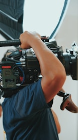 A person operates a large professional camera, holding it on their shoulder. The camera is equipped with various controls, cables, and a matte box. Strong lighting is visible in the background, indicating a studio or professional filming environment.