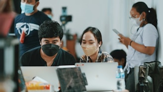 Several people are gathered in an indoor setting, with two individuals in the foreground focused on laptops while wearing masks. Others are standing in the background, with one person using a smartphone. The scene appears to be collaborative, possibly in a work or educational environment.