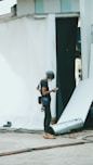 A person wearing a helmet and safety gear is standing in an industrial or construction-like setting. They are looking at a mobile device, possibly for work-related purposes. Around them are sheets or fabric drapes hanging, with cables and construction materials scattered on the concrete ground.
