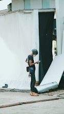 A person wearing a helmet and safety gear is standing in an industrial or construction-like setting. They are looking at a mobile device, possibly for work-related purposes. Around them are sheets or fabric drapes hanging, with cables and construction materials scattered on the concrete ground.