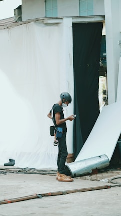 A person wearing a helmet and safety gear is standing in an industrial or construction-like setting. They are looking at a mobile device, possibly for work-related purposes. Around them are sheets or fabric drapes hanging, with cables and construction materials scattered on the concrete ground.