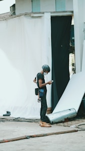 A person wearing a helmet and safety gear is standing in an industrial or construction-like setting. They are looking at a mobile device, possibly for work-related purposes. Around them are sheets or fabric drapes hanging, with cables and construction materials scattered on the concrete ground.