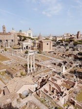 A panoramic view of ancient ruins featuring columns, arches, and fragmented structures. There are multiple levels of archaeological excavations, surrounding greenery, and modern city buildings in the background.