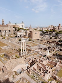 A panoramic view of ancient ruins featuring columns, arches, and fragmented structures. There are multiple levels of archaeological excavations, surrounding greenery, and modern city buildings in the background.