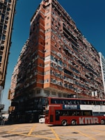 A large, multi-story residential building with a worn, red exterior dominates the scene. Numerous windows and air conditioning units are visible on the building's facade, and the ground level contains various shop signs and commercial businesses. In the foreground, a red double-decker bus with advertisements is crossing a yellow-box junction on a sunny day.