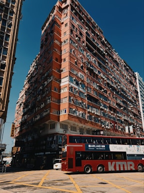 A large, multi-story residential building with a worn, red exterior dominates the scene. Numerous windows and air conditioning units are visible on the building's facade, and the ground level contains various shop signs and commercial businesses. In the foreground, a red double-decker bus with advertisements is crossing a yellow-box junction on a sunny day.
