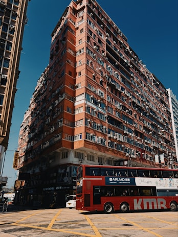 A large, multi-story residential building with a worn, red exterior dominates the scene. Numerous windows and air conditioning units are visible on the building's facade, and the ground level contains various shop signs and commercial businesses. In the foreground, a red double-decker bus with advertisements is crossing a yellow-box junction on a sunny day.
