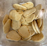 A clear plastic container filled with small, oval-shaped pieces of flatbread. The bread has a light brown, toasted appearance and appears soft and fluffy, with small air pockets visible on the surface. The container is positioned on a light, neutral background.