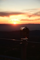 A silhouette of a stationary viewing binocular against the backdrop of a vibrant sunset. The sky is filled with warm hues of orange, red, and yellow as the sun sets on the horizon, with clouds scattered above. The binoculars are positioned in the foreground, casting a dark shadow, and a metal fence or railing can be seen.
