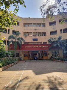 A multi-story building with a clear sign indicating it is the Faculty of Architecture & Ekistics. The entrance is adorned with plants and trees, providing a natural, welcoming atmosphere. The architecture features geometric lines and large windows on its facade. Two people are visible at the entrance, adding a sense of scale to the building.