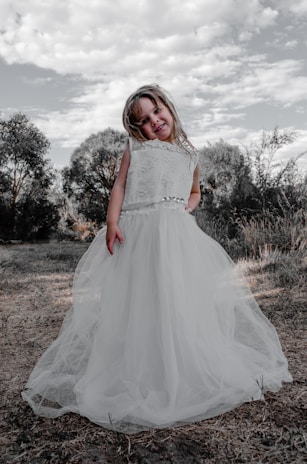 A joyful girl wearing a flowing white dress with floral patterns, standing in a sunlit garden.