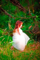 A child with long, auburn hair is wearing a white dress while standing in a lush, green forest. The scene is vibrant, with dense foliage and a fallen tree branch creating a natural, whimsical atmosphere.