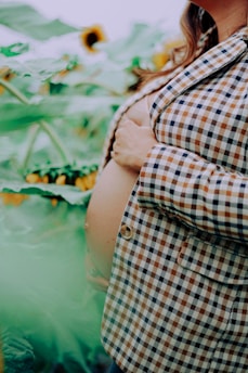 A pregnant woman wearing a plaid jacket is gently holding her belly. She is standing in a field of sunflowers, with large green leaves surrounding her.