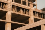 A partially constructed concrete building with exposed steel reinforcements and wooden scaffolding. The unfinished structure displays multiple open floors and columns, creating a grid pattern. Electrical wires are visible in the foreground against a clear sky.