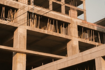 A partially constructed concrete building with exposed steel reinforcements and wooden scaffolding. The unfinished structure displays multiple open floors and columns, creating a grid pattern. Electrical wires are visible in the foreground against a clear sky.