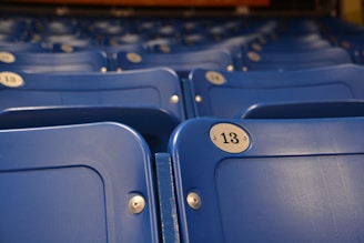 Rows of empty blue plastic seats are lined up, each with a circular metal number plate. The seat in the foreground is labeled with the number 13. The focus is on the seats, with a shallow depth of field blurring the background.