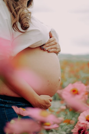 A pregnant woman gently cradles her belly with her hand while standing in a field of vibrant pink flowers. Her brown hair cascades over a white shirt and she is wearing blue jeans. The foreground and background are filled with soft-focus floral elements, creating a serene atmosphere.