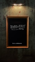 A blackboard encased in a wooden frame is mounted on a grungy, industrial-style wall with a single overhead light fixture illuminating it. The word 'KHOO-KAM' and some additional text are written on the blackboard.