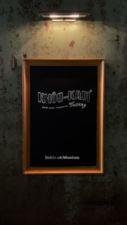 A blackboard encased in a wooden frame is mounted on a grungy, industrial-style wall with a single overhead light fixture illuminating it. The word 'KHOO-KAM' and some additional text are written on the blackboard.